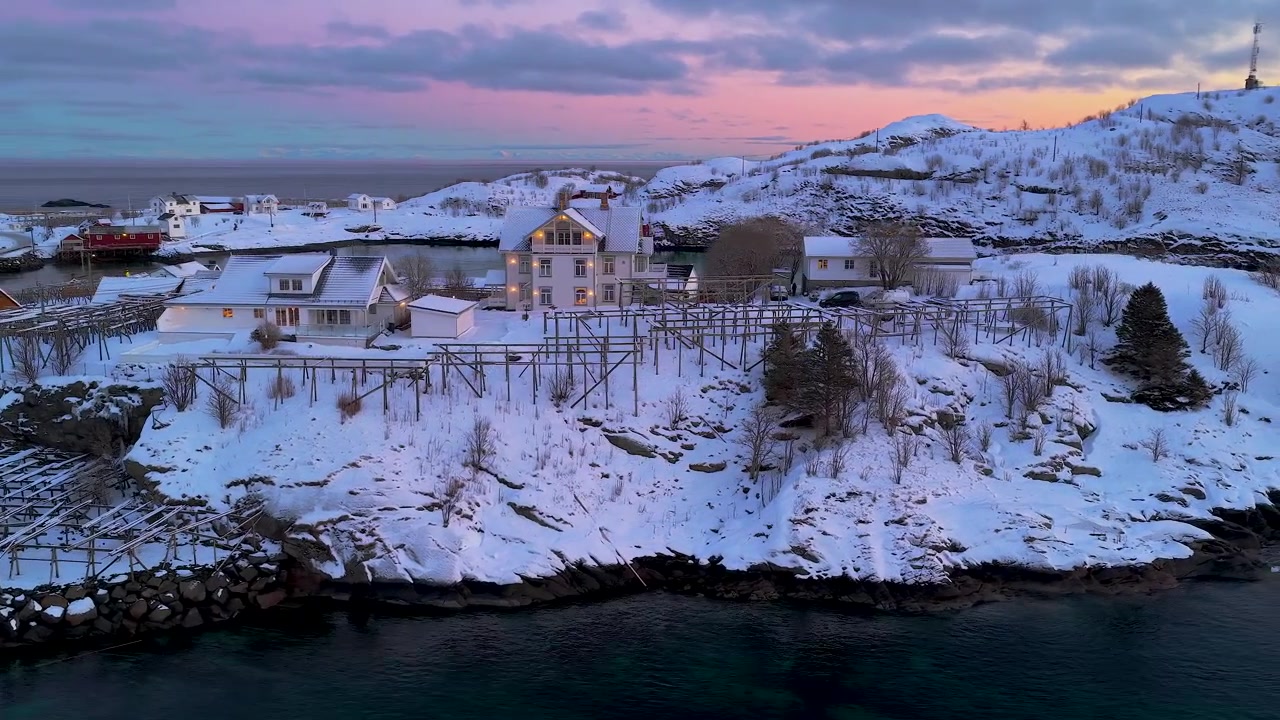 ゴールデンアワーのノルウェー、ロフォーテン諸島にある雪に覆われた漁村のシネマティックな空撮。ピンク色の夕焼け空を背景に、伝統的な家屋と魚の干し棚が特徴。