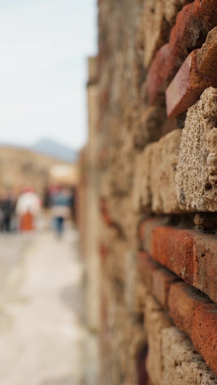 Vertical close-up of weathered Pompeii brick masonry with shallow depth of field, while tourists and the ancient street fade softly into the background.