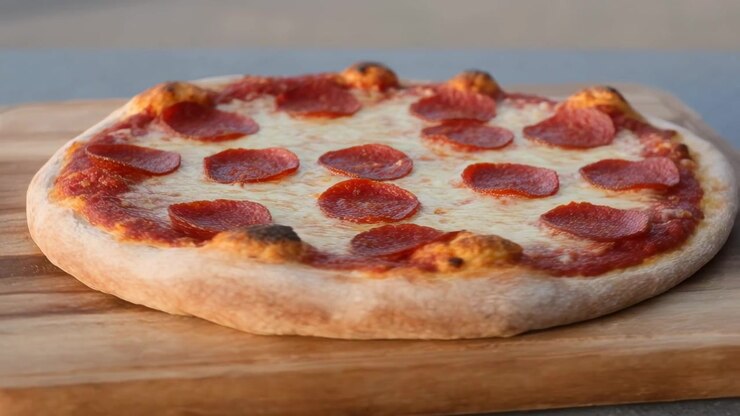 La telecamera si allontana e si alza, rivelando una pizza ai peperoni su un tagliere di legno, mostrando la sua forma rotonda completa.