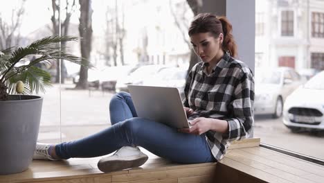 hermosa mujer morena trabajando en su computadora portátil. sentada cerca de la ventana en un café con interior moderno. fondo borroso de la calle