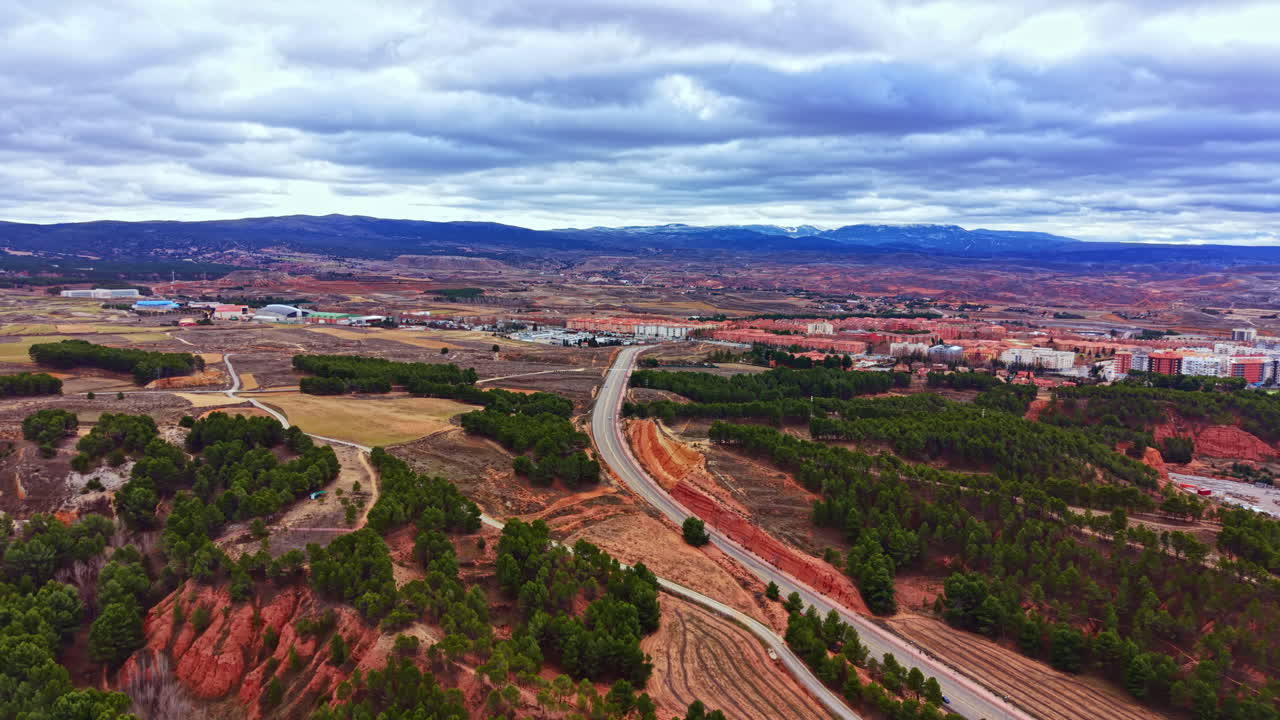 白天，西班牙小鎮附近，多雲的天空下，鄉村景觀中一條蜿蜒的道路穿梭於山丘和樹木之間。