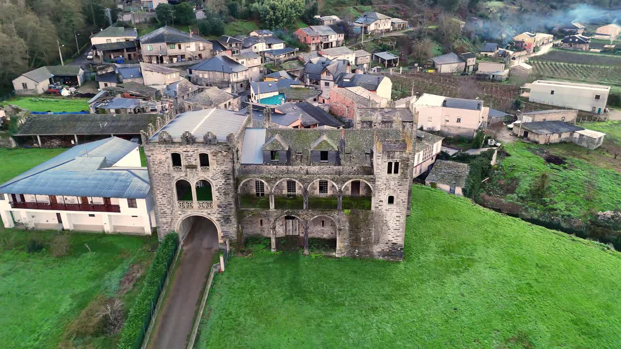 Aerial shot of Arnedo Castle, a historic stone fortress with arches and towers, surrounded by green fields and a small village in Valdeorras.