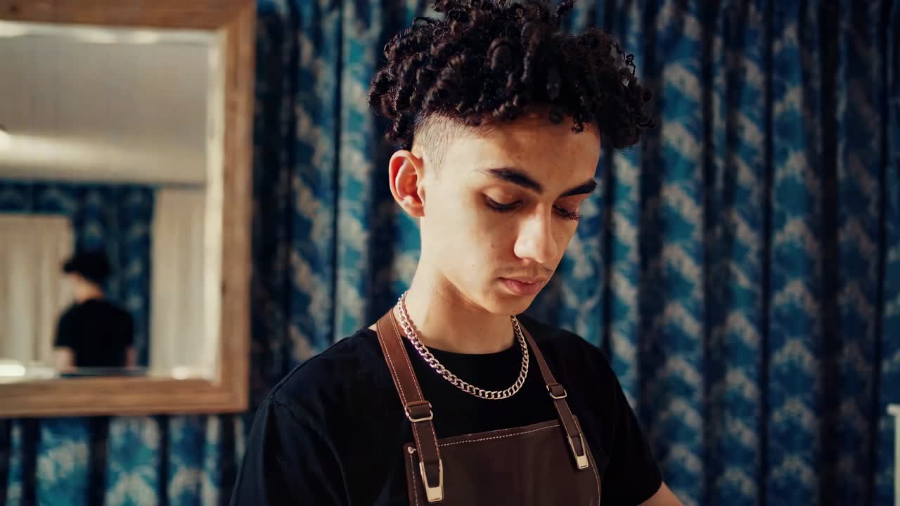 Young male barber wearing an apron and looking downwards with concentration while working in his vintage salon with blue curtains and a mirror