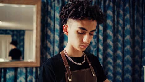 young male barber wearing an apron and looking downwards with concentration while working in his vintage salon with blue curtains and a mirror