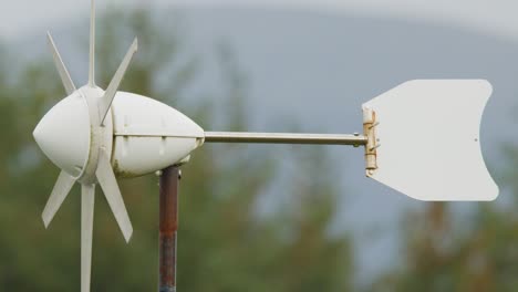 a white wind turbine spins rapidly on a metal pole outdoors, with blurred trees and sky in the background. soft daylight, steady camera, side view