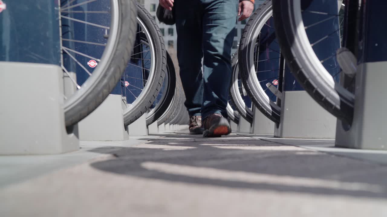 A man with orange top with a black hoodie walks in between the parked public bike tires towards the camera in a sunny day.