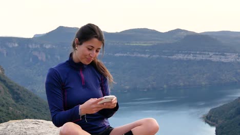 cheerful young ethnic woman smiling and using smartphone on rocky cliff