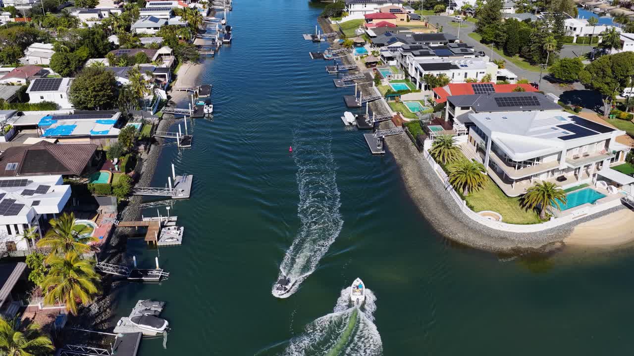 A speedboat travels along a residential canal lined with luxury homes in Gold Coast, Australia, captured in bright daylight with smooth aerial camera movement