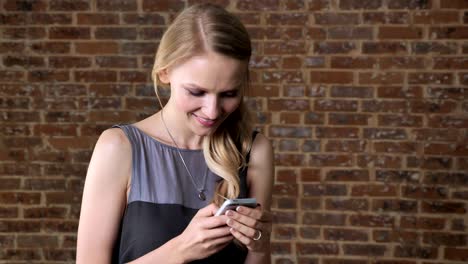 young cute blond girl is watching photos on smartphone, showing it, smiling, communication concept, brick background