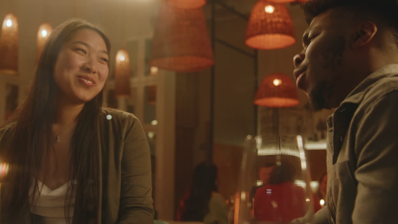 Multi Ethnic Couple Sit and Talk in Cafe African American Man and Asian Woman Sit in Modern Gastro Cafe together Talk Multi Ethnic Couple on a Date in Restaurant Waiting for Order the Glass with Wine on Table Concept of Public Catering