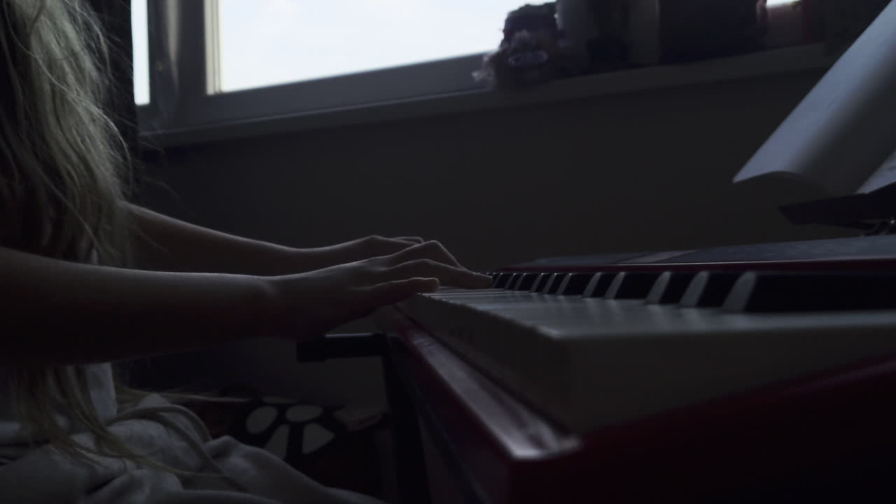 A moody, static side-angle shot capturing a young girl's hands as she plays a red digital piano. The footage focuses on the coordination between both hands on the keys, set in a quiet, indoor environm