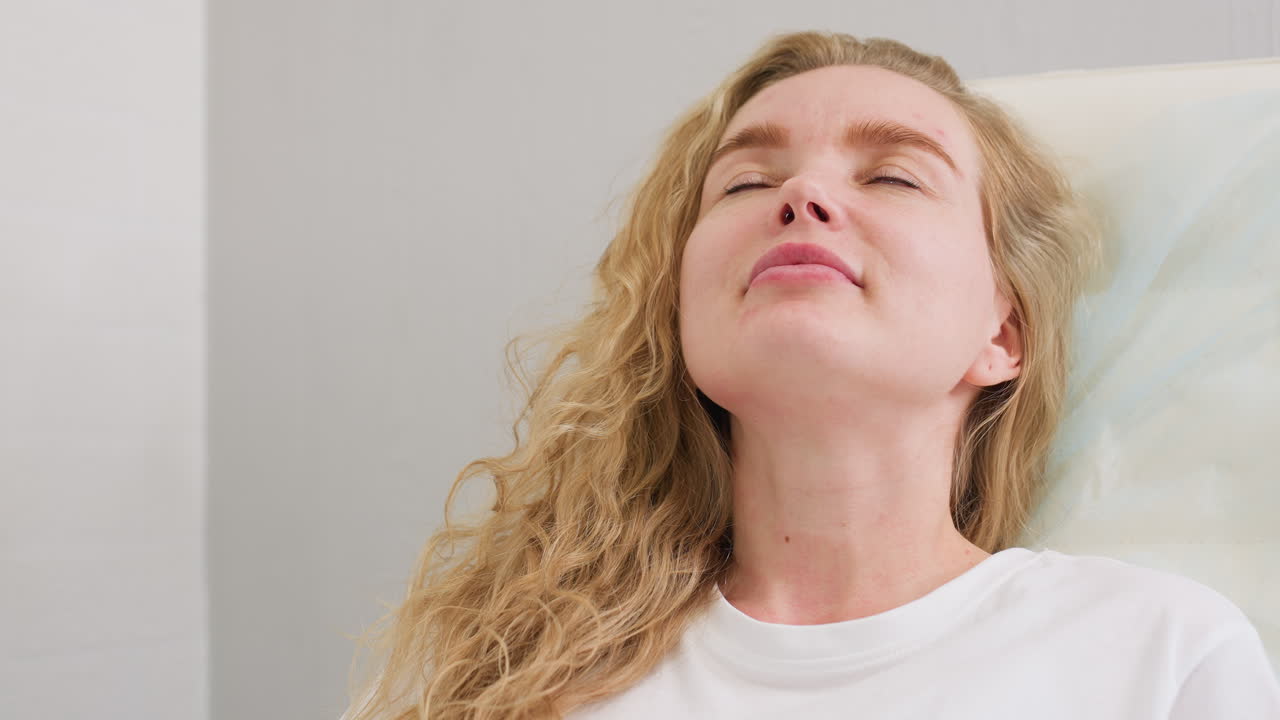Blond client in white polo shirt lying back with head on chair, looking calm and composed during beauty or wellness session in clean indoor setting, conveying relaxation and self-care
