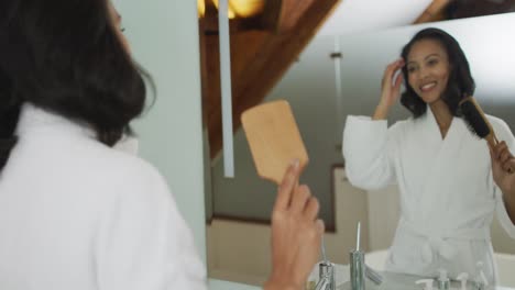 Mixed-race-woman-wearing-bathrobe-looking-at-mirror-and-brushing-her-hair