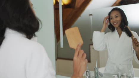 mixed race woman wearing bathrobe looking at mirror and brushing her hair