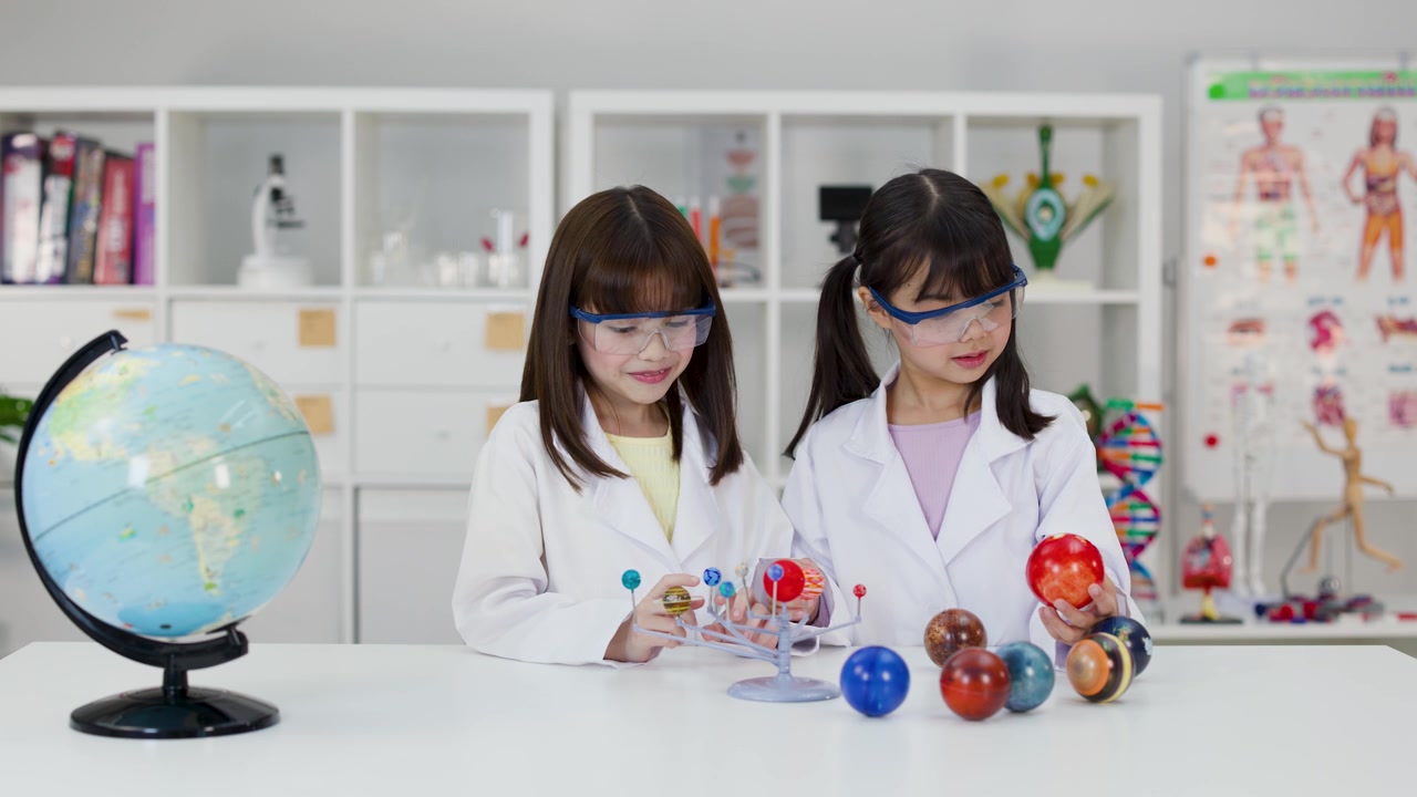 Two young students wear lab coats and goggles while examining a solar system model together.