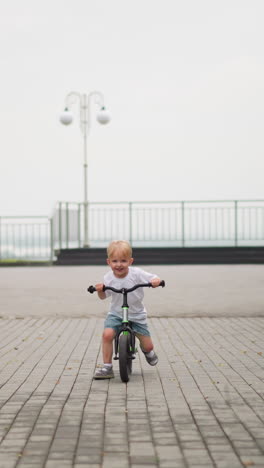 a happy little boy rides his bicycle down a paved path with a big smile, in the blurred background, other children can be seen walking
