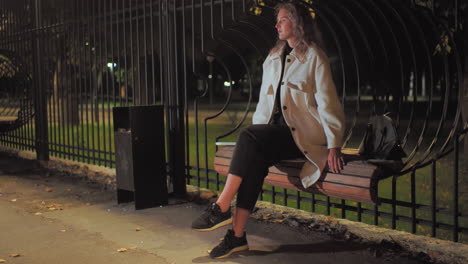 young lady wearing white coat and black pants sits calmly on wooden bench near iron fence at night, relaxing under soft street lights in peaceful park environment with fallen leaves around