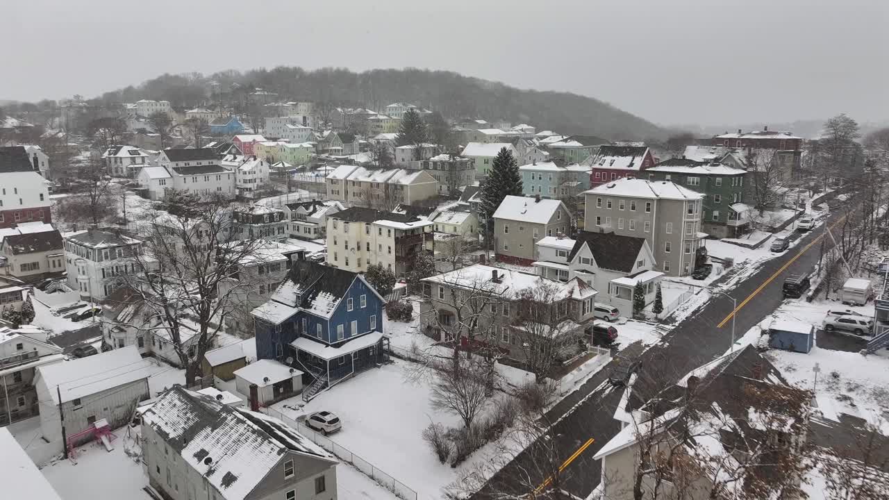 Snow-covered houses in a New England neighborhood. Colorful homes and bare trees on hilly terrain. Peaceful aerial of small-town winter charm.