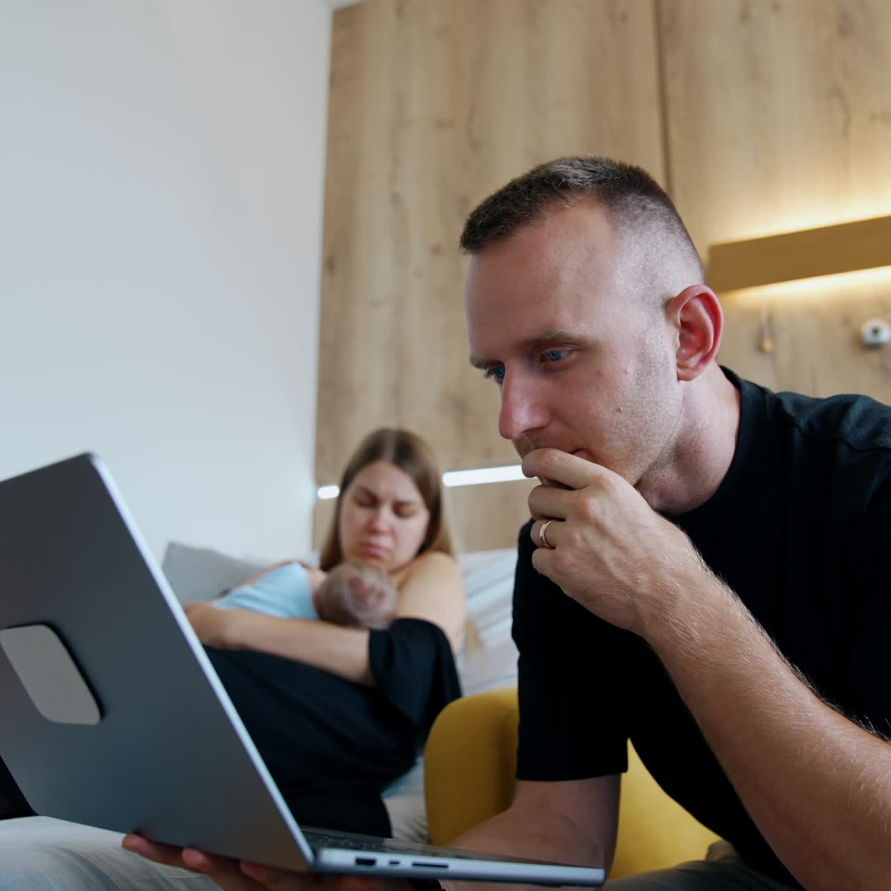 Long-haired woman sitting on the bed is focused on breastfeeding a newborn. Man sits beside in the armchair focused on laptop