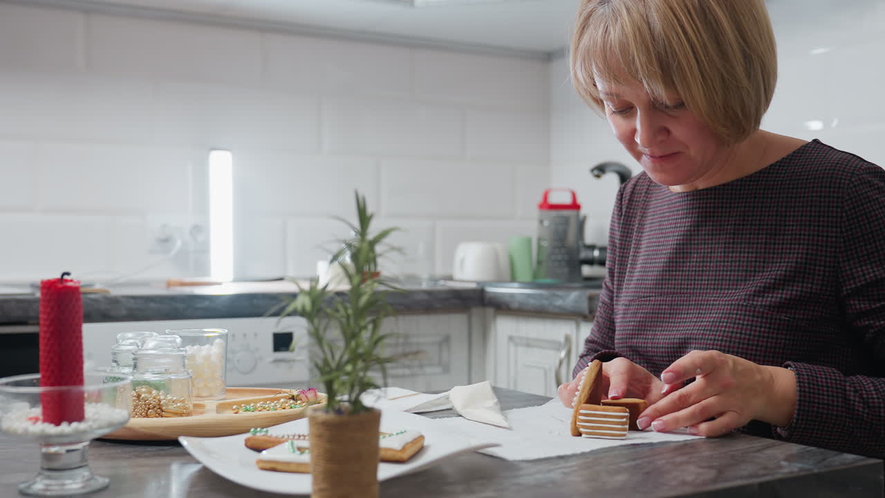 Woman using cookies to design a house, surrounded by flower-shaped cookies, baking essentials, and decorative beads on kitchen table, creating festive cookies in a cozy kitchen atmosphere