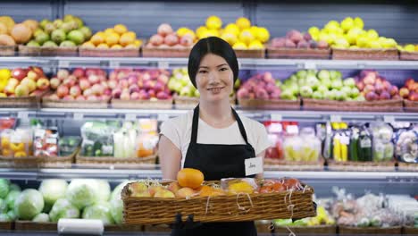 Retrato-De-Una-Atractiva-Joven-Trabajadora-Asiática-Parada-En-Un-Supermercado-Con-Estantes-De-Frutas-En-El-Fondo,-Mirando-La-Cámara-Y-Sonriendo.-Concepto-De-Negocio-Y-Personas-De-Comercio