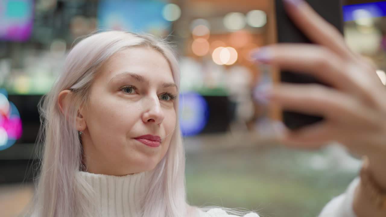 Cheerful mall visitor sits beside water fountain on bench adjusting hair with chic manicure and posing with mobile phone, capturing sparkling jets under bright mall lights while shoppers pass by