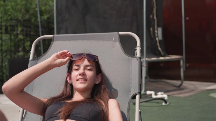 Young woman relaxes on gray lounge chair outdoors adjusting her sunglasses and lying back. Woman wears black swimsuit in bright sunlight with greenery and wooden structures visible