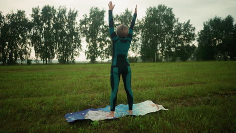 back view of woman in green and black suit and leggings doing yoga on mat in grassy field, arms raised in peaceful motion as she moves slowly, surrounded by vast open field and trees in the distance