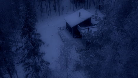 cozy small cabin at night in snow covered forest, aerial drone view