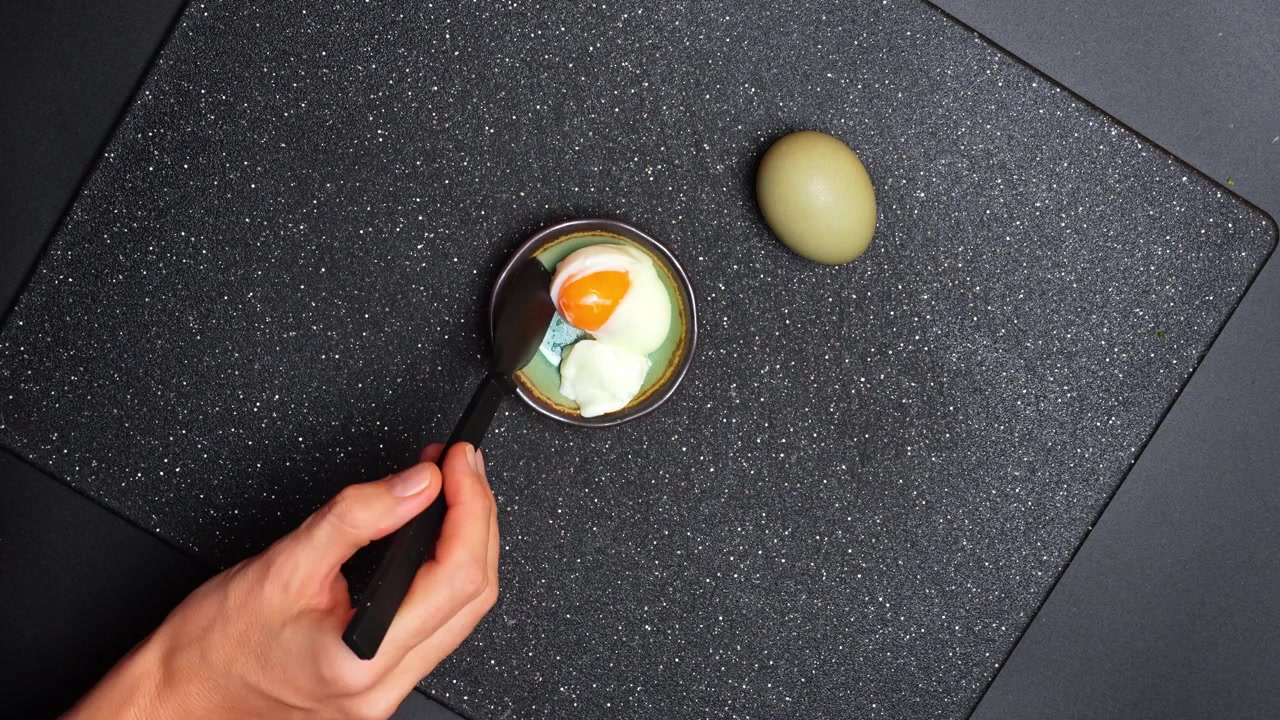 Person peeling a soft boiled egg over a bowl. Top view of hands removing the shell from a pale green egg over a small ceramic dish.