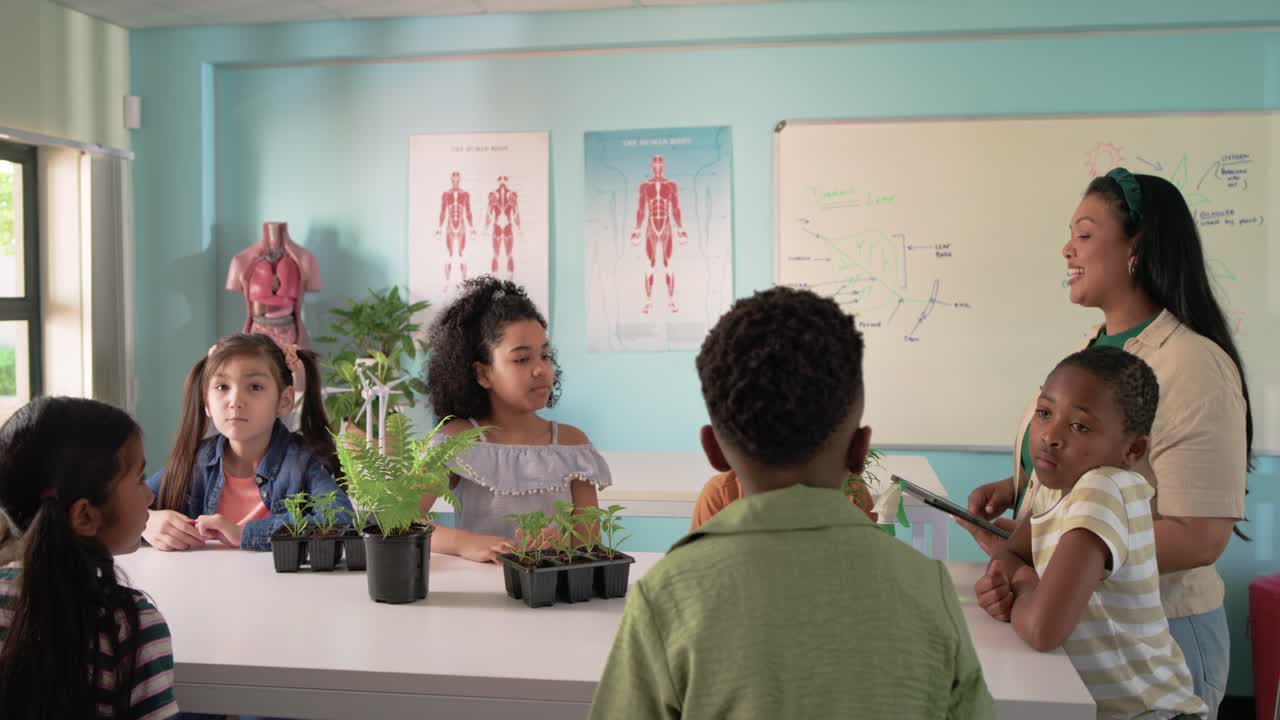 African American teacher using tablet urging kids examining plants with lens and spray in class