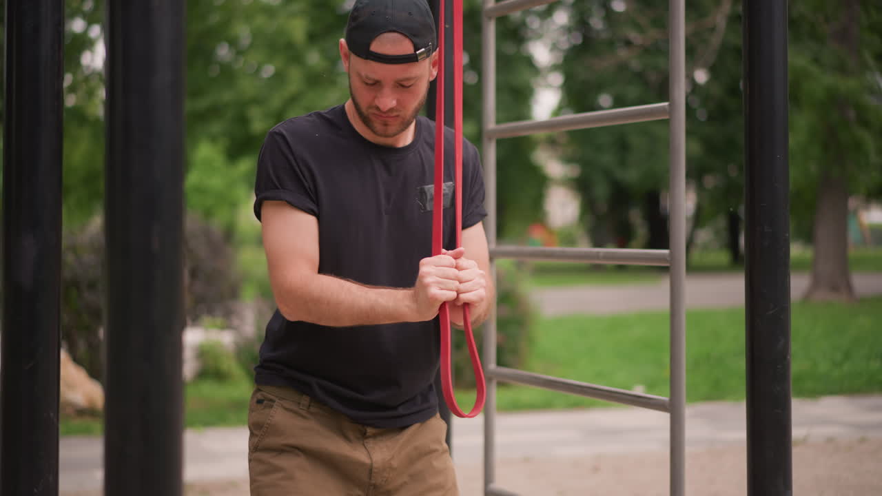 Man Adjusting Outdoor Climbing Rope, Person Creating Tension With Red Cord On Gym Apparatus Backdrop, Male Figure Focused On Securing Red Rope Around Outdoor Fitness Equipment In Park Setting