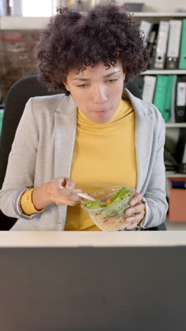 biracial businesswoman uses laptop, eats lunch in slow-motion vertical video
