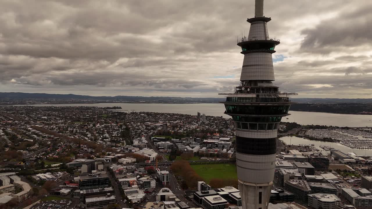 Aerial orbit of sky tower with marina port and cityscape of Auckland city, New Zealand. Wide shot. Cloudy day in summer. Suburb neighborhood in distance