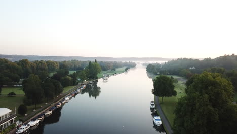 beautiful drone shot of henley on thames at sunrise, flying along the river to the east with a light mist over the river and surrounding hills and fields