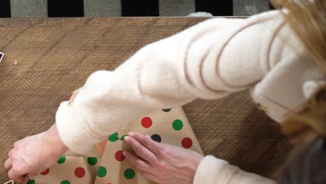 una mujer joven con un abrigo de lana se esfuerza por envolver un regalo de navidad con papel de envoltura de puntos decorativos y cinta adhesiva en una mesa de madera