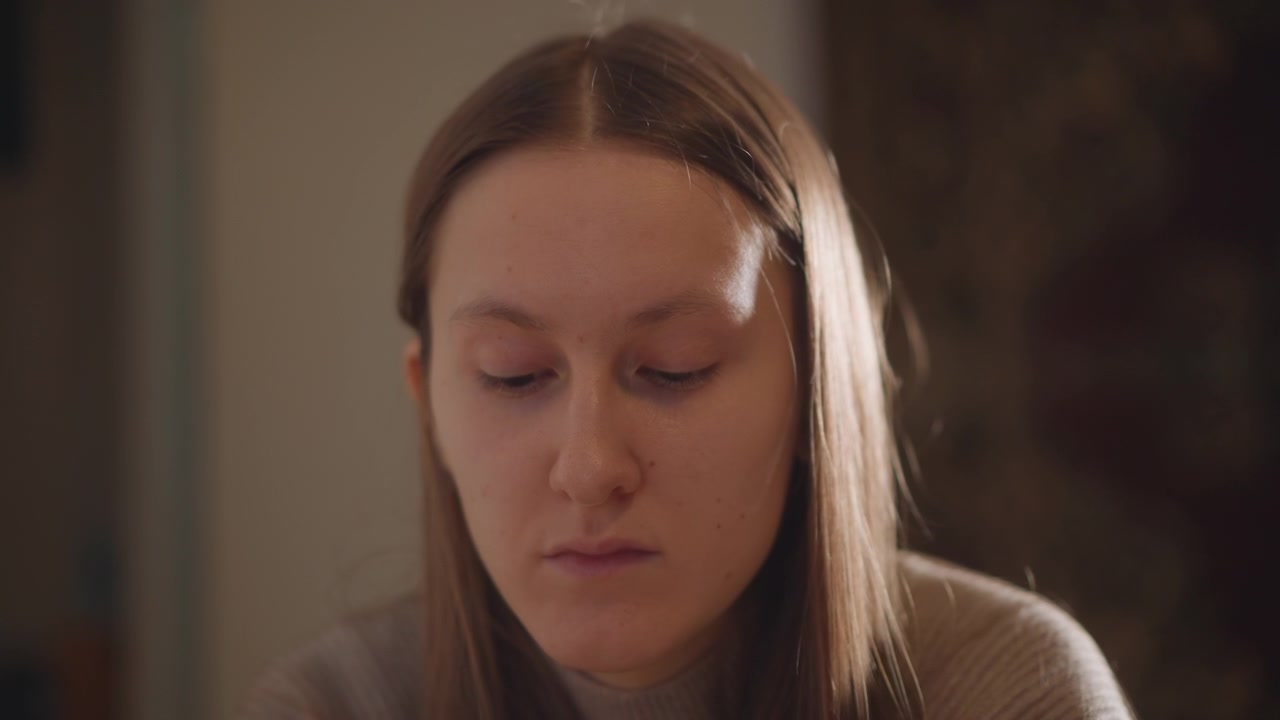 Young woman sitting contemplative indoors, soft cinematic light on face, closeup portrait of student journaling and reflecting at evening desk, subtle melancholy expression, warm lamp glow, textured