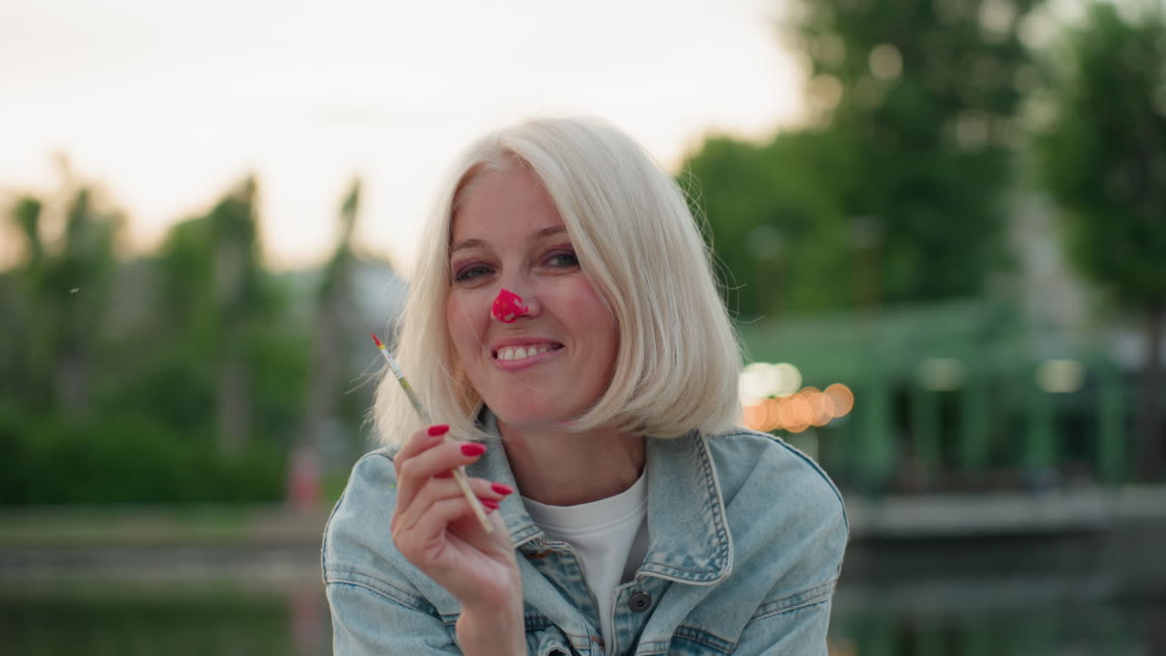 portrait of cheerful mom holding paint brush, bright pink paint on nose, smiling warmly during playful lakeside painting session at sunset, paint splatters on denim jacket and joyful expression