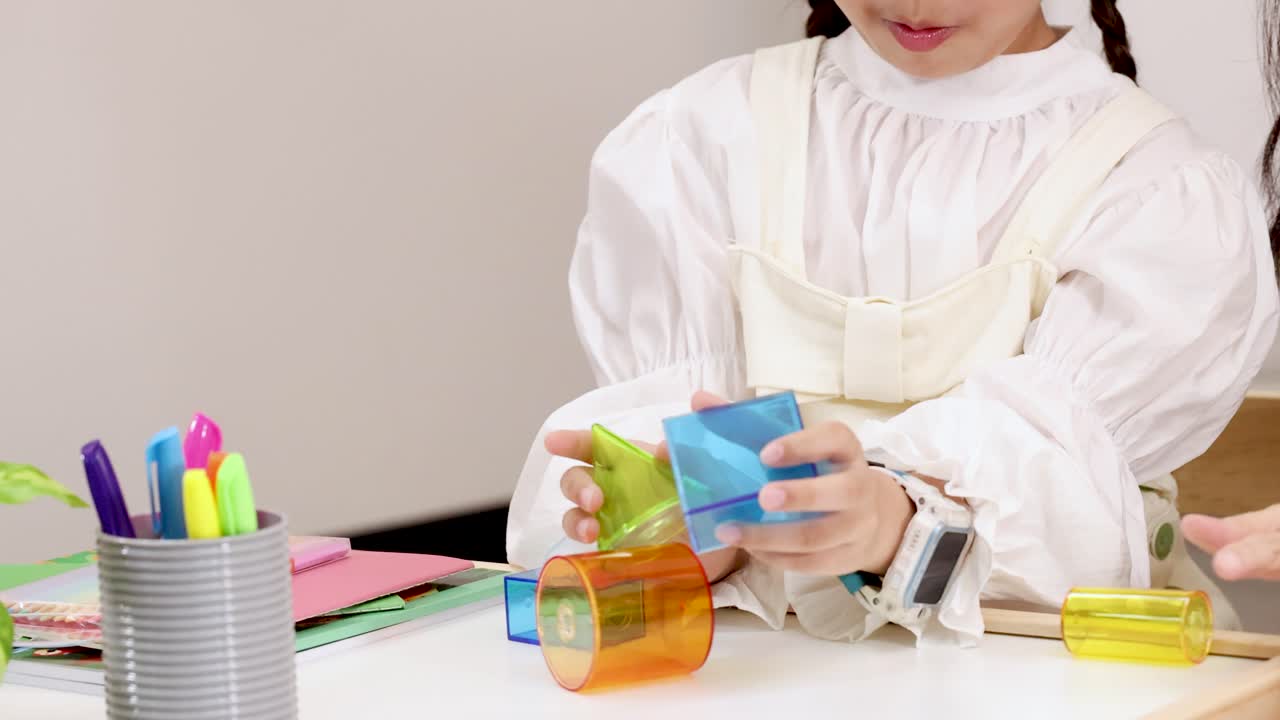 Young girl builds tower with translucent geometric blocks, which tumbles onto white desk, bright lighting