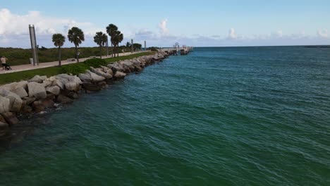 Water-flyover-along-beach-pier-with-long-jetti-and-palm-trees-into-atlantic-ocean