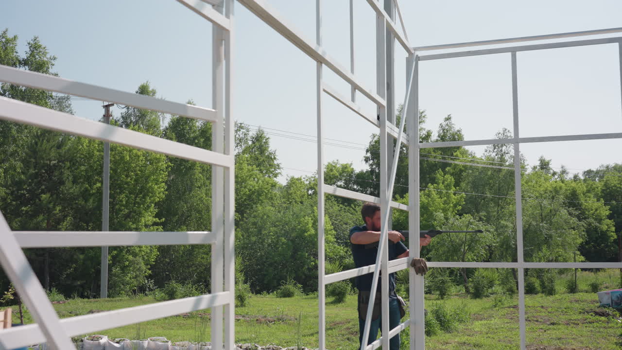 man aiming rifle at sky near greenhouse frame to scare birds from garden while standing among plants under bright sun, movement focused on protection of crops
