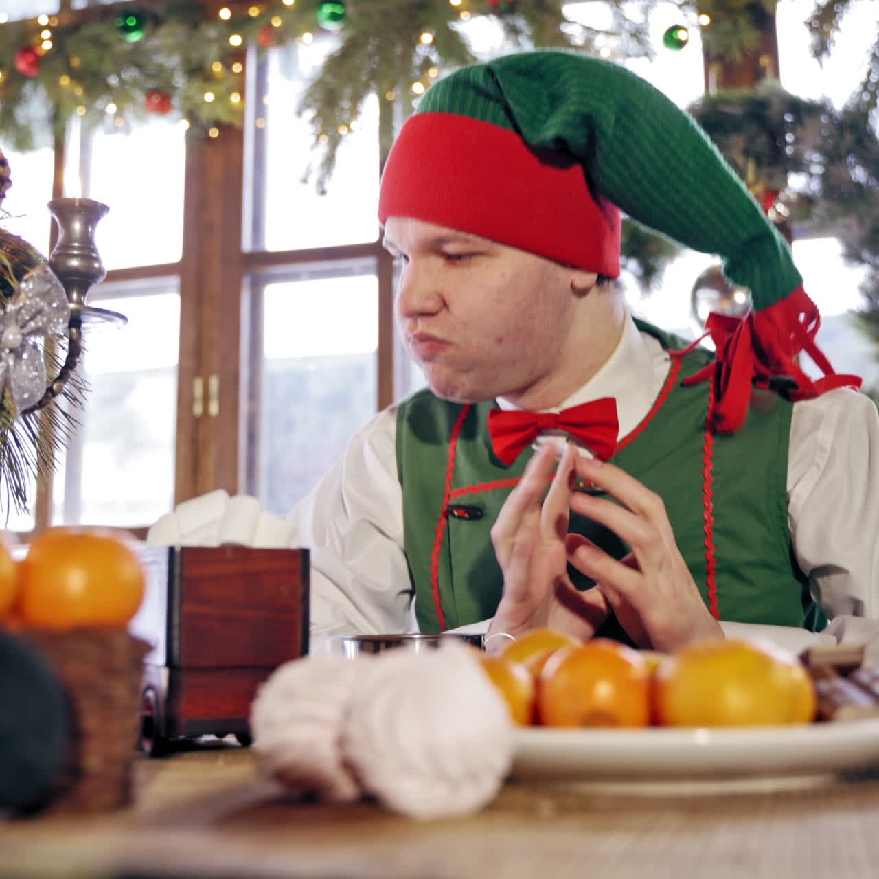 Hungry elf drinking at the table at Christmas. Male elf in green costume with hood eating at festive table on decorated background.