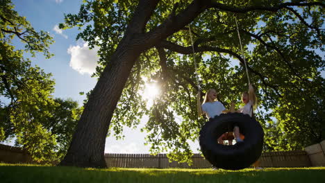 two girls playing on a tire swing under a large tree