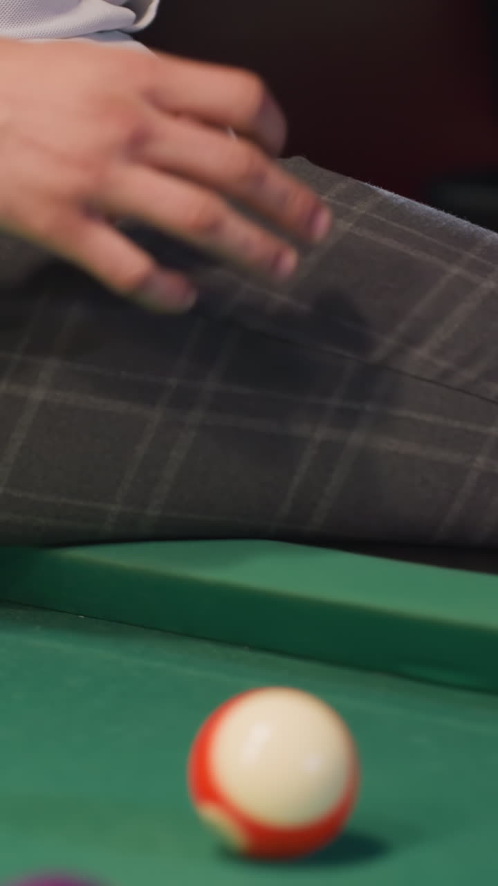 Close-up of person in white shirt and grey trousers sitting on green pool table, rolling billiard ball. Focus on lower body, stylish attire, and casual indoor ambiance. Relaxed setting