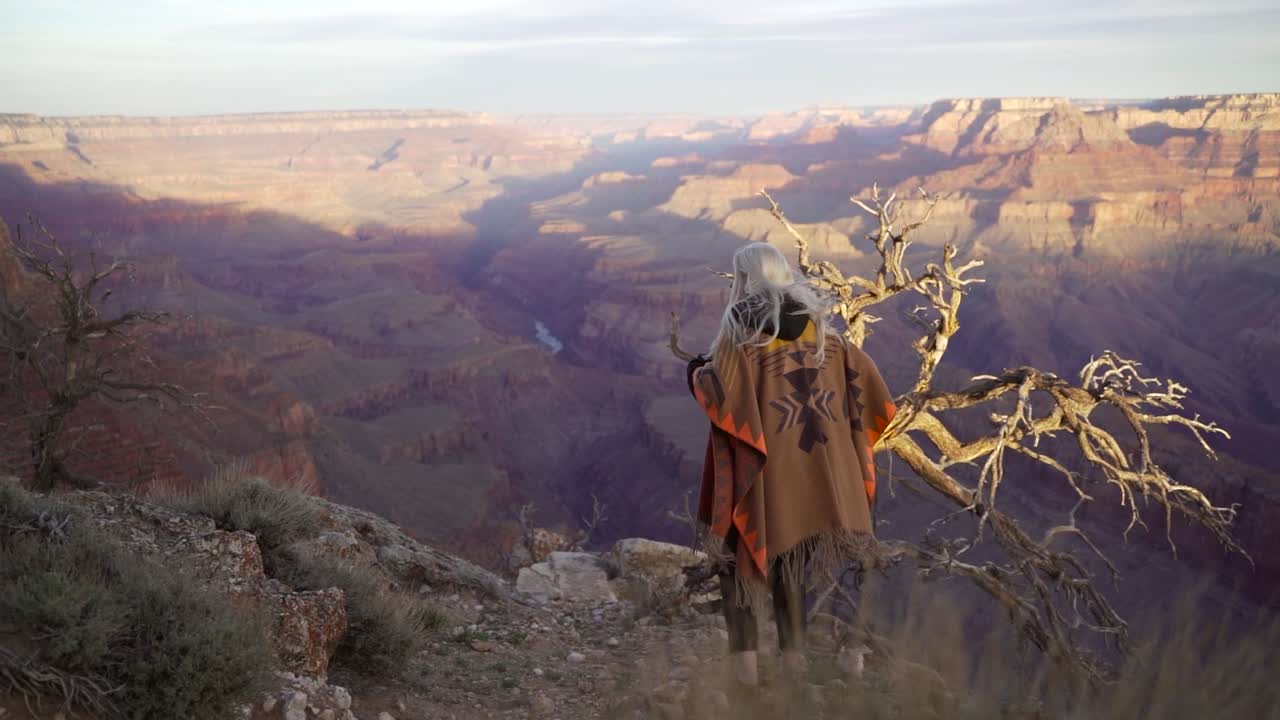 전통적인 패턴과 금발 머리카락을 가진 갈색 오렌지색 담요를 입은 관광 하이커 소녀의 뒷면은 가장자리 절벽과 그랜드 캐니언을 내려다보는 파노라마 전망을 볼 수 있습니다.