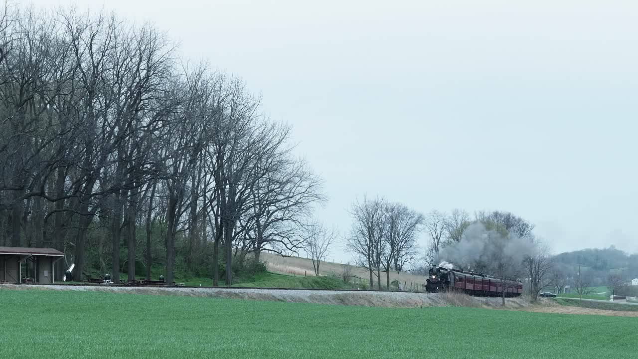 A vintage steam train chugs along a rural track, emitting dark smoke as it moves past a small shelter surrounded by trees. The tranquil landscape reflects evening ambiance.