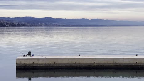 Fisherman-on-peer-on-Sea-shore-with-town-in-distance-close-up,-Opatija,-Croatia