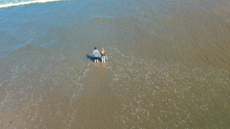 toma aérea reveladora de una pareja de pie de la mano en el océano en la playa de noventa millas con olas rodando sobre la playa, nueva zelanda