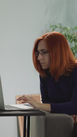 redhead freelancer puts on glasses to protect eyes while working on laptop from home. woman sits on sofa in living room looking in electronic device