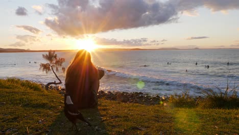 Mujer-Sentada-En-La-Orilla-Del-Mar-Cubierta-De-Hierba-Y-Turistas-Surfeando-En-La-Playa-En-El-Parque-Nacional-De-Noosa-Al-Atardecer